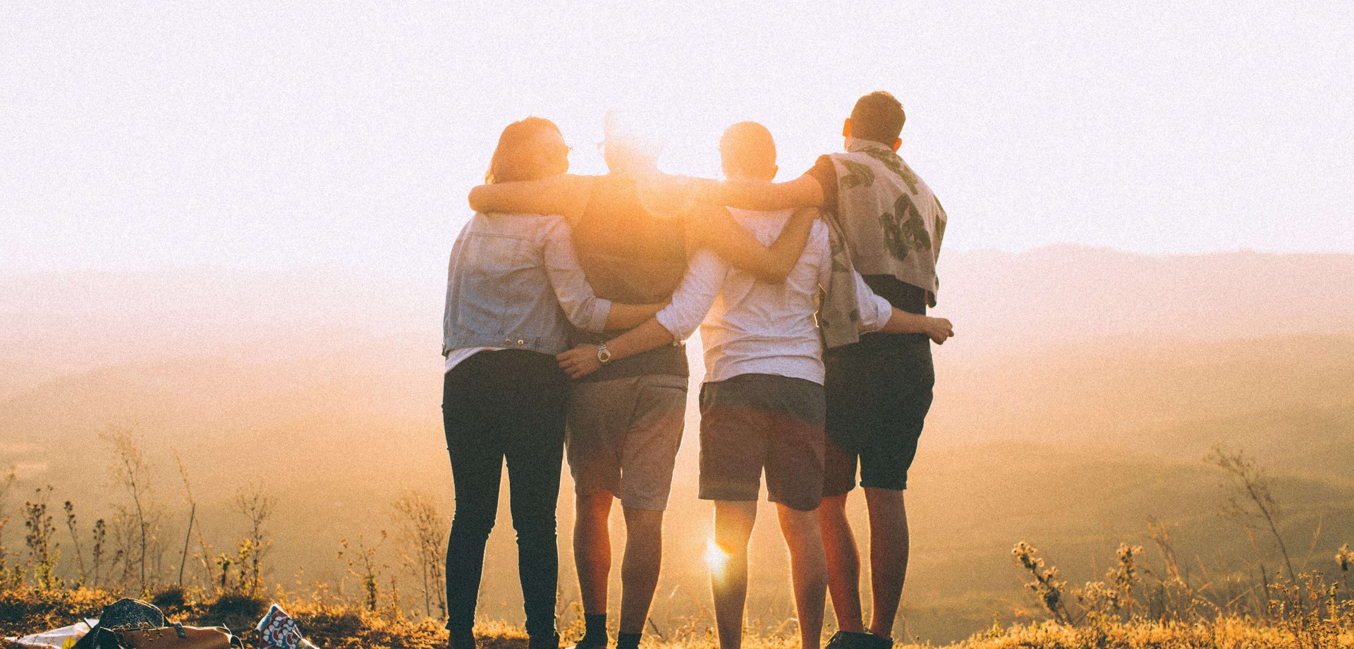 Das Bild zeigt vier junge Menschen, die auf einem Berg stehen und mit dem Rücken zum Betrachtenden Arm in Arm in die Sonne schauen. ©Pexels / Helena